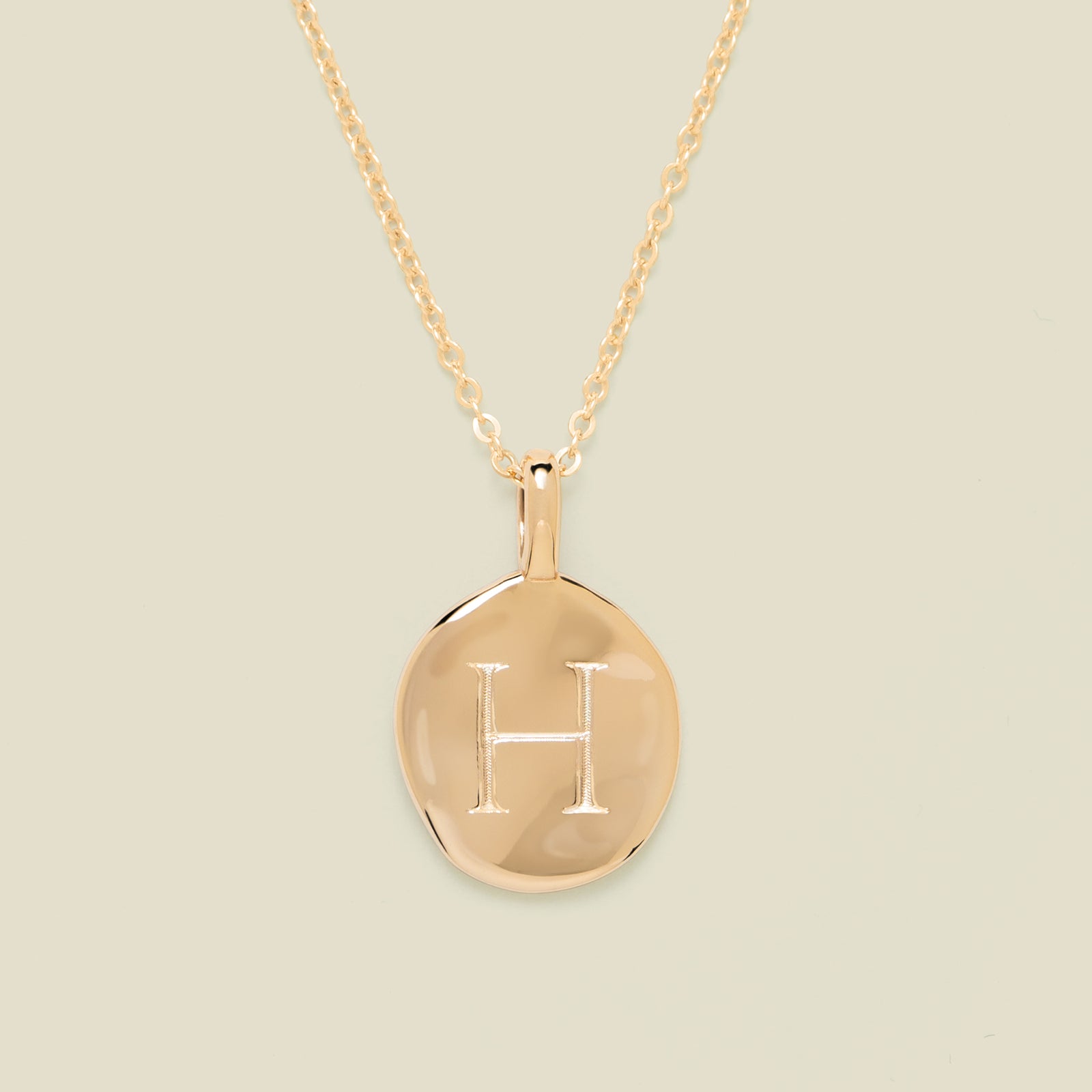 Gold necklace with a round pendant engraved with the letter 'H' on a beige background