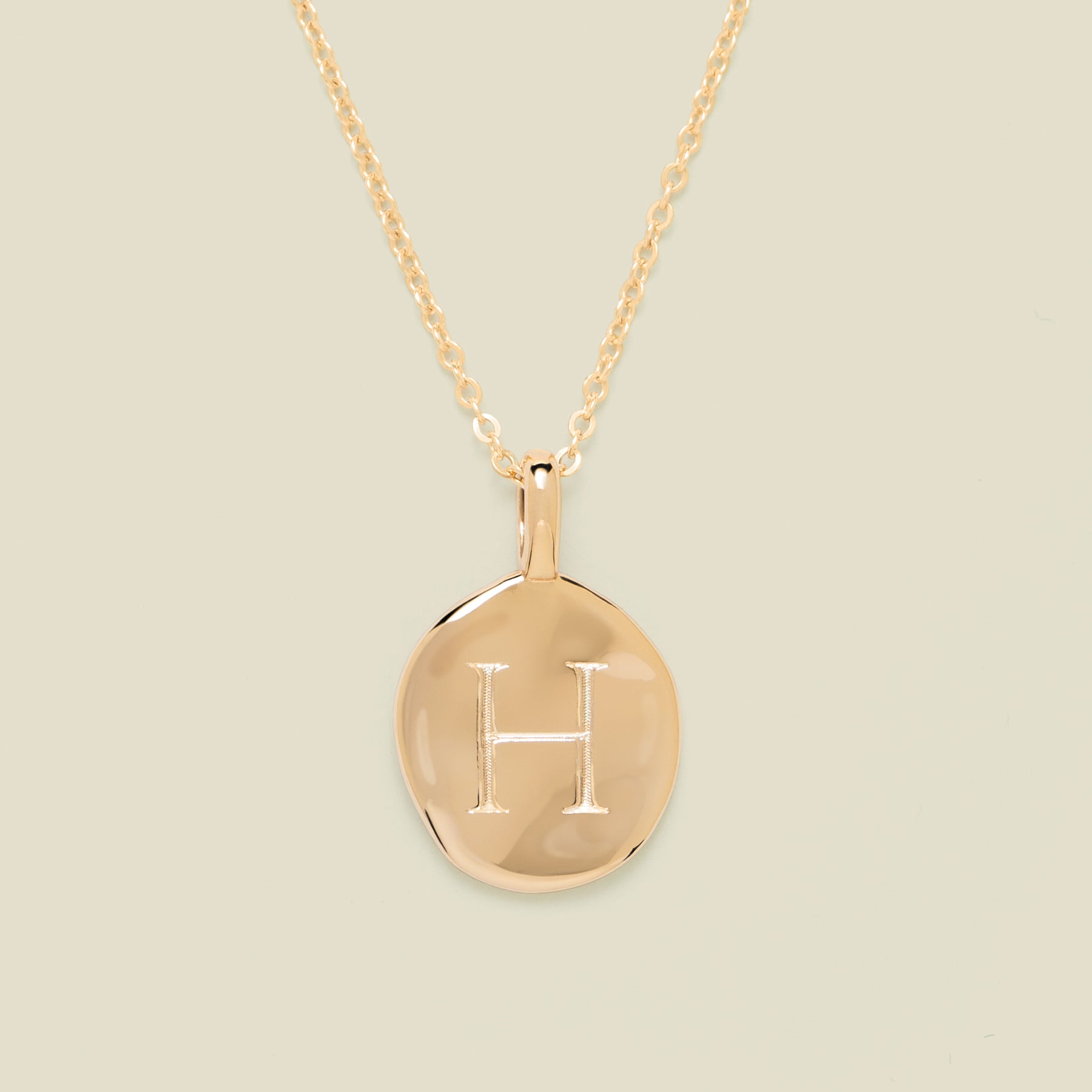 Gold necklace with a round pendant engraved with the letter 'H' on a beige background