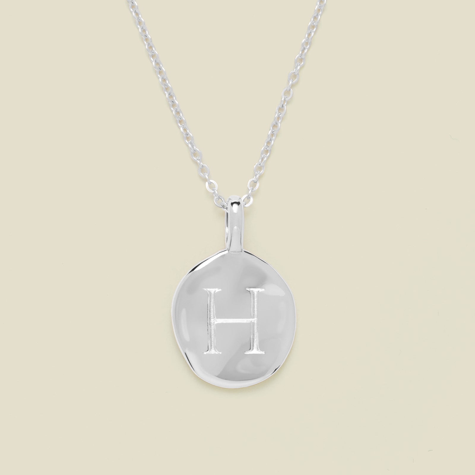 Silver necklace with a round pendant engraved with the letter 'H' on a beige background