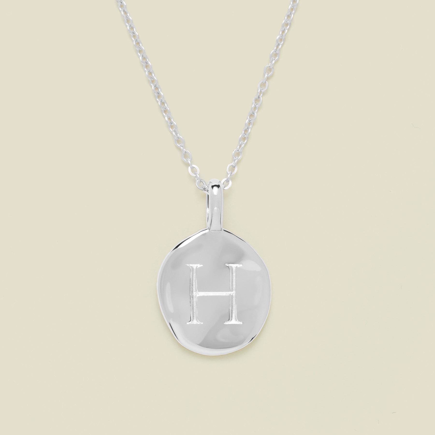 Silver necklace with a round pendant engraved with the letter 'H' on a beige background