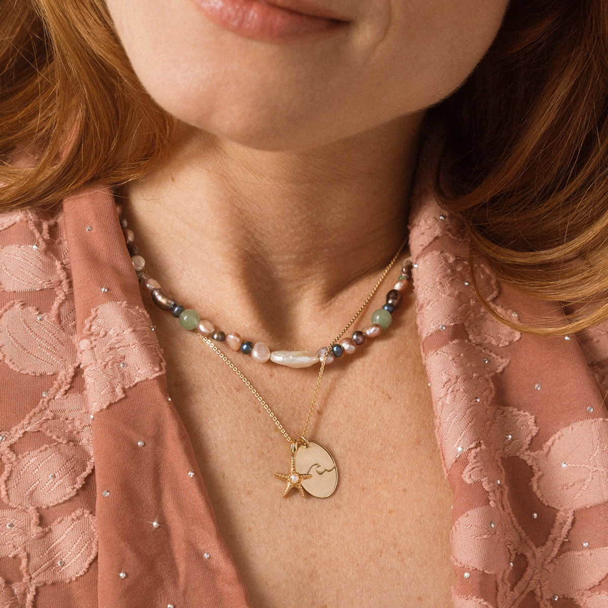 Close-up of a woman's neck wearing a  gold necklace with a starfish charm paired with a multicolored pearl necklace. 