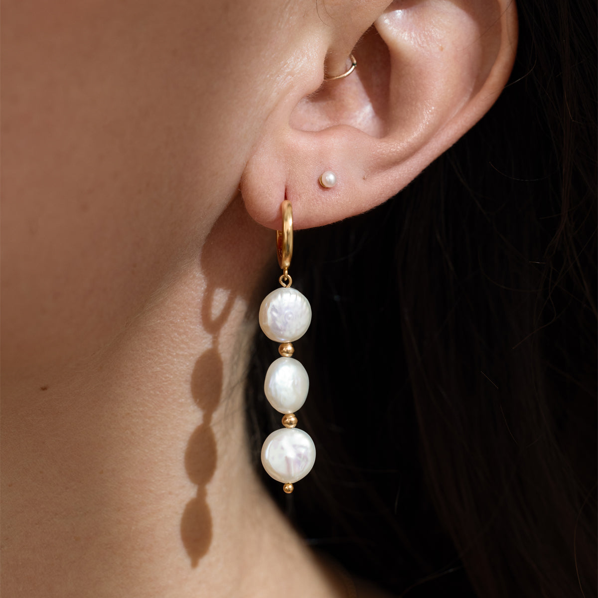 Close-up of an ear wearing a gold hoop earring with three dangling white pearls, paired with a small pearl stud above.