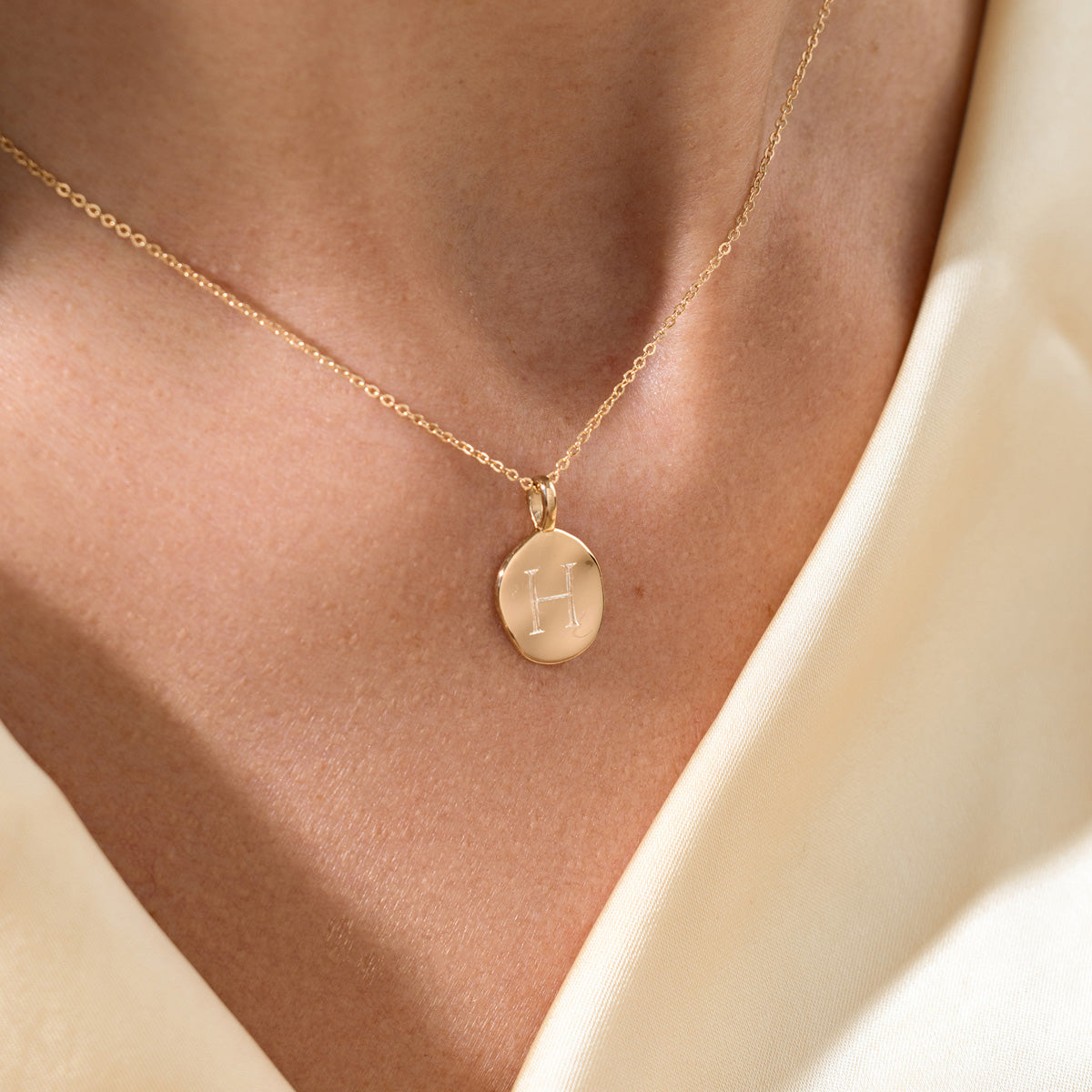 Gold necklace with a pendant engraved with the letter 'H' on a person wearing a beige top.