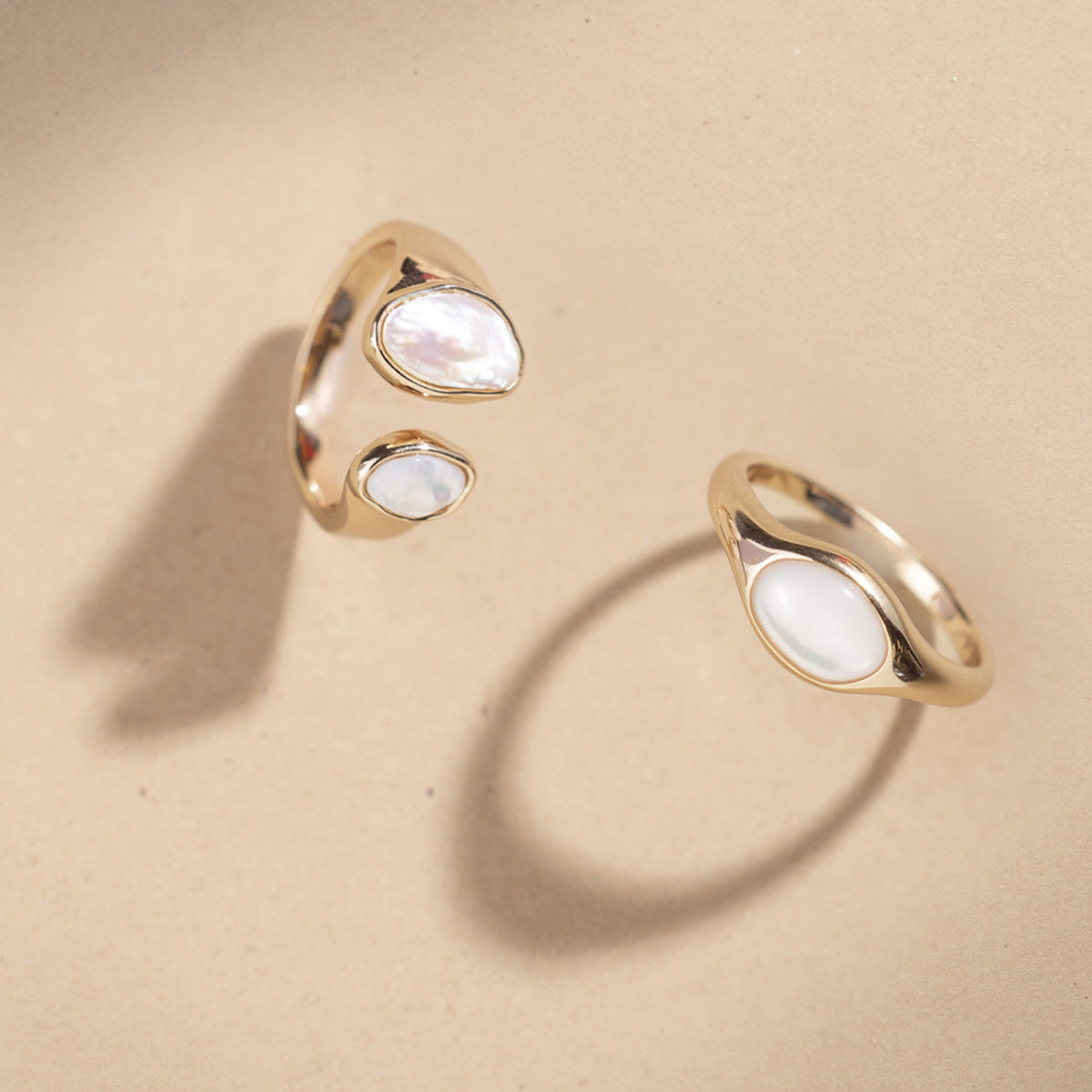 Two gold bezel-set pearl rings against a plain beige background.