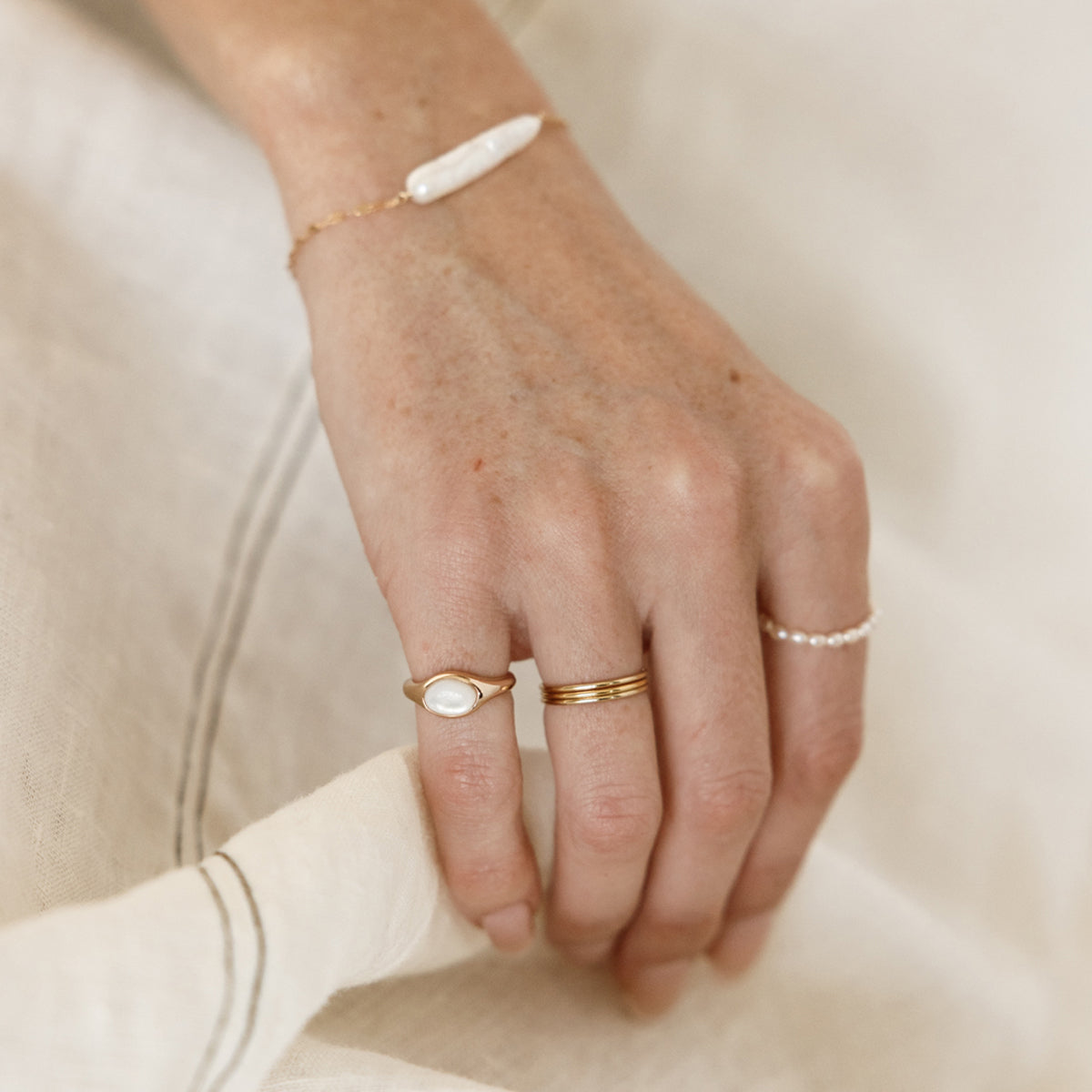 Hand lightly touching linen wearing gold stacked rings, a bezel-set pearl pinky ring and a baroque pearl bracelet.