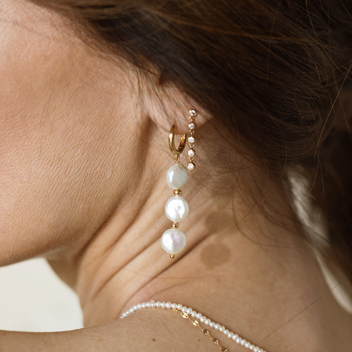 Close-up of an ear wearing a gold hoop earring with three dangling pearls, paired with a delicate row of small gemstone studs and a fine pearl necklace visible along the shoulder.