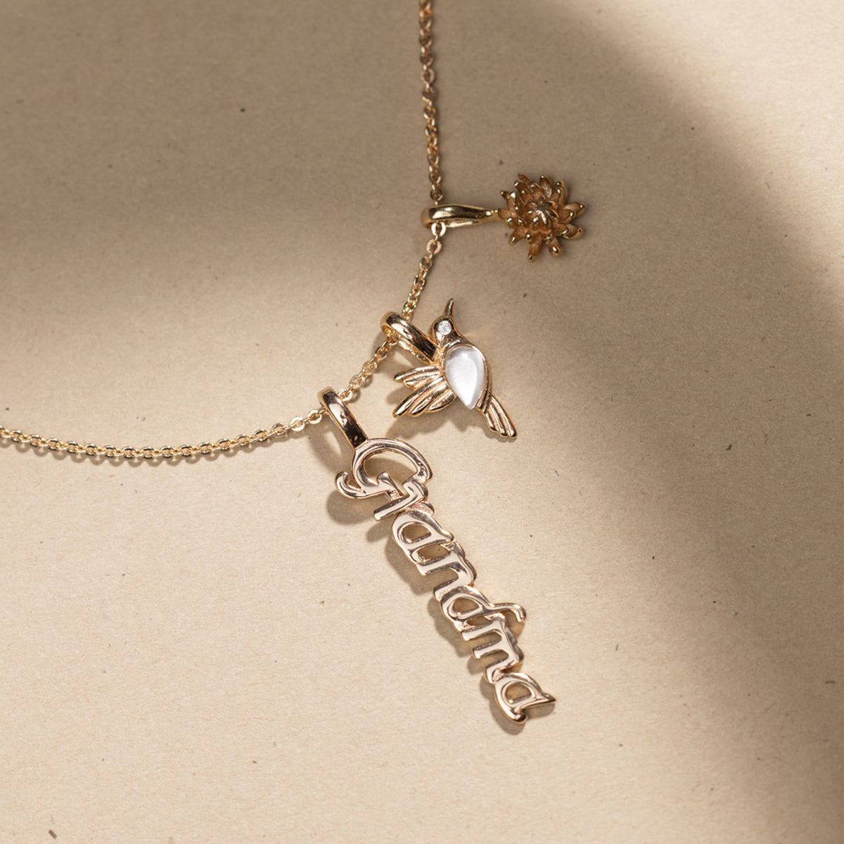 Gold necklace with 'Grandma', hummingbird and Chrysanthemum charm pendants on a beige background